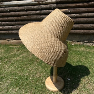 Puede incluir: Un sombrero de paja de ala ancha de color marr&oacute;n y textura tejida. El sombrero se muestra en un soporte de madera.