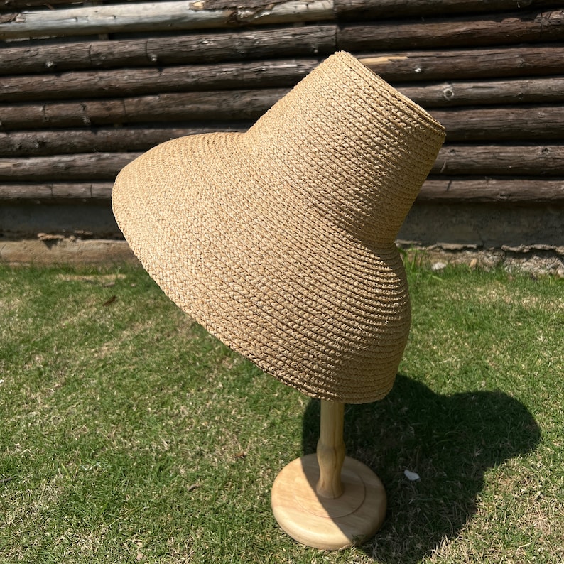 Puede incluir: Un sombrero de paja de ala ancha de color marr&oacute;n y textura tejida. El sombrero se muestra en un soporte de madera.