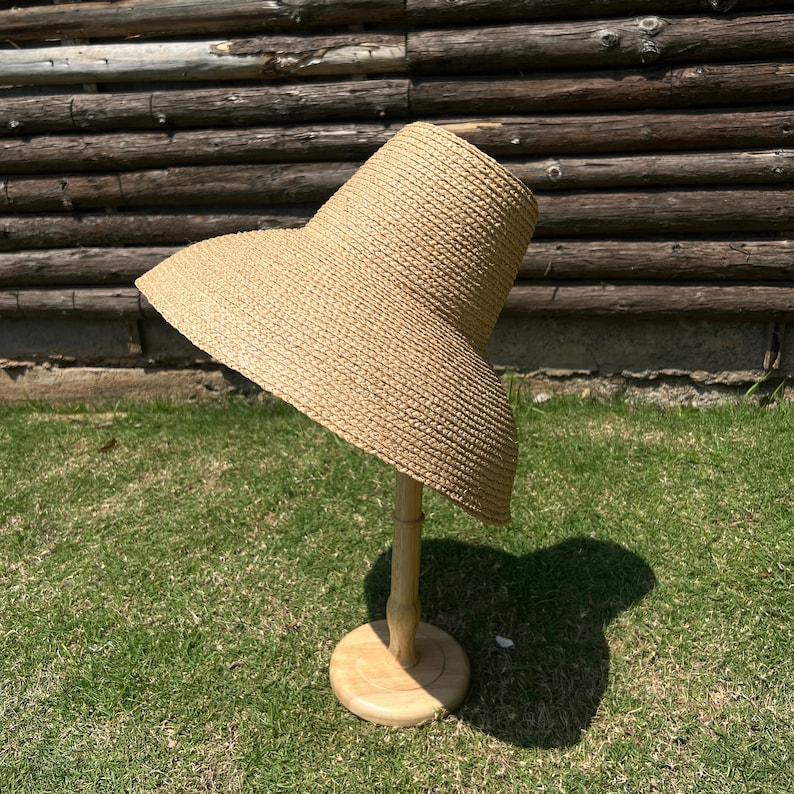 Puede incluir: Un sombrero de paja de ala ancha de color marr&oacute;n claro, exhibido en un soporte de madera. El sombrero tiene una textura tejida y una corona ligeramente redondeada.