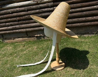 May include: A large, wide-brimmed straw hat with a tall, pointed crown. The hat has a white ribbon tied around the crown and long white ribbons hanging down from the brim.