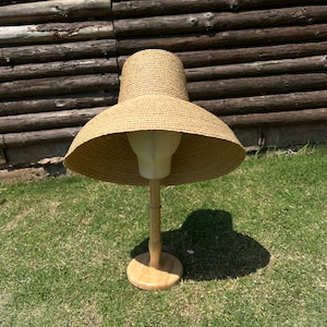 Puede incluir: Un sombrero de paja de ala ancha de color marr&oacute;n, exhibido en un soporte de madera. El sombrero tiene una ala ancha y plana y una corona ligeramente redondeada.