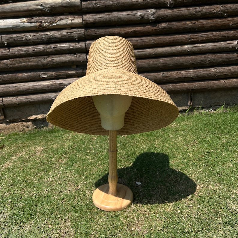 Puede incluir: Un sombrero de paja de ala ancha de color marr&oacute;n, exhibido en un soporte de madera. El sombrero tiene una ala ancha y plana y una corona ligeramente redondeada.