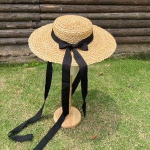 Puede incluir: Un sombrero de paja de ala ancha con una cinta negra atada en un lazo. La cinta se extiende en largas ataduras que fluyen.