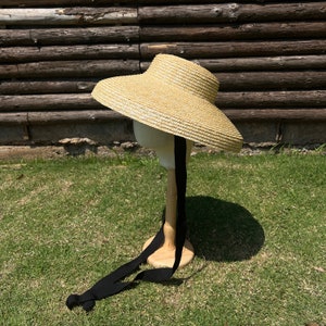 May include: A wide-brimmed straw hat with a black ribbon tied around the crown. The hat is sitting on a wooden stand against a wooden background.