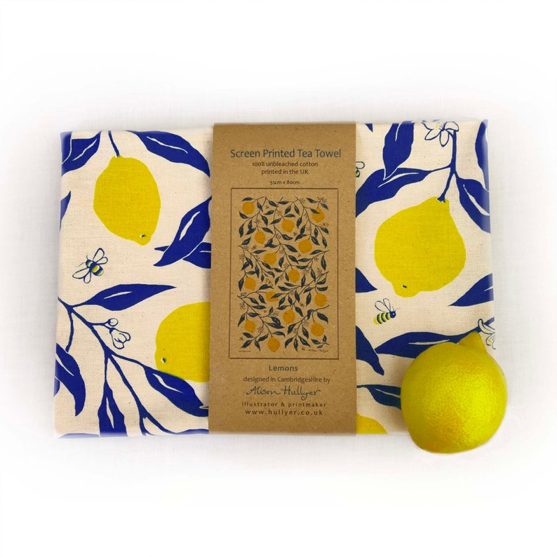 May include: A tea towel with a lemon print. The towel is white with blue leaves and yellow lemons. The text on the tag reads "Screen Printed Tea Towel 100% unbleached cotton printed in the UK SunxBoom Lemons designed in Cambridgeshire by Alison Hullyer illustrator & printmaker www.hullyer.co.uk"