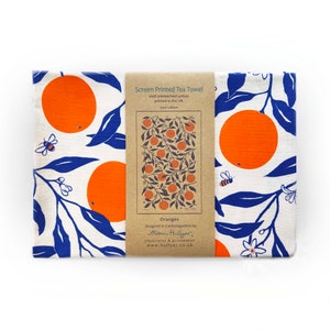 May include: A white tea towel with a blue and orange orange tree design. The towel is screen printed with 100% unbleached cotton and measures 5cm by 80cm. The text on the label reads "Screen Printed Tea Towel", "100% unbleached cotton", "printed in the UK", "5cm x 80cm", "Oranges", "designed in Cambridgeshire by Alison Hullyer", "Illustrator & printmaker", "www.hullyer.co.uk".