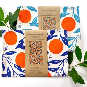 May include: Three tea towels with a white background and a blue and orange orange tree design. The towels are packaged with a brown paper label that says "Screen Printed Tea Towel" and "Oranges".