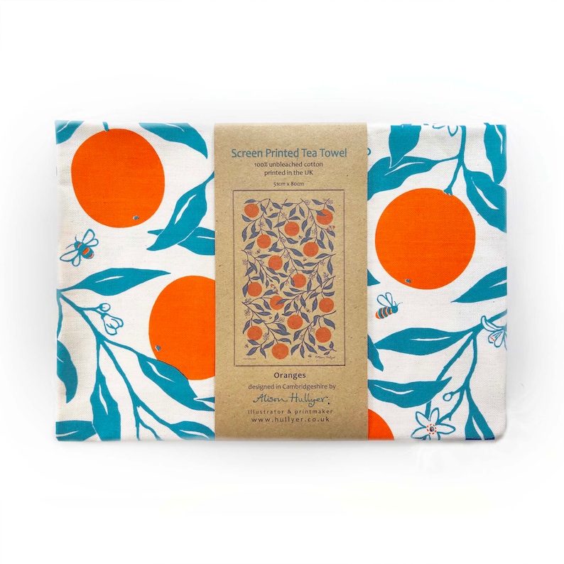 May include: A white tea towel with a screen printed design of oranges on branches with blue leaves. The towel is packaged with a brown paper label that reads "Screen Printed Tea Towel 100% unbleached cotton printed in the UK 5cm x 80cm Oranges designed in Cambridgeshire by Alison Hullyer, Illustrator & printmaker www.hullyer.co.uk"