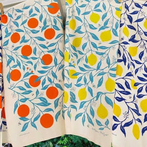 May include: Four tea towels with a white background and a repeating pattern of blue leaves, white flowers, and either orange or yellow citrus fruit. The text "Atom Hullyer" is visible on the bottom right towel.