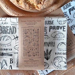 May include: Three white tea towels with black illustrations of different types of bread and baking terms. The towels are folded and displayed on a wooden surface. The text on the towels includes "Rise", "Prove", "Loaf", "It Pain", "Ciabatta", "Banary", "Soda Brea", "Knead", "Croissant", "Catch Rolls", "Split Tin", "Bread", "Crusty Loaf", "Stoneground", "Grissini", "Rye", "Wh", "Baking", "Designed in Cambridgeshire by", "Aloon Hullyer", "Illustrator & Printmaker", "www.hullyer.co.uk".