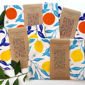 May include: Five tea towels with a white background and a repeating pattern of blue leaves, white flowers, and either yellow lemons or orange oranges. The towels are folded and arranged on a white surface with green leaves. Each towel has a brown paper tag with the text "Screen Printed Tea Towel" and the name of the fruit.