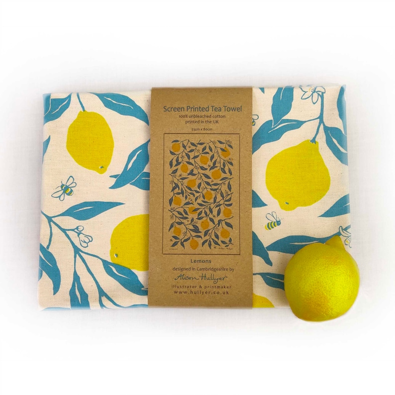 May include: A tea towel with a lemon print. The towel is made of 100% unbleached cotton and is printed in the UK. The towel is 30cm x 30cm. The text on the label reads "Screen Printed Tea Towel 100% unbleached cotton printed in the UK 30cm x 30cm Lemons designed in Cambridgeshire by Alison Hullyer illustrator & printmaker www.hullyer.co.uk"
