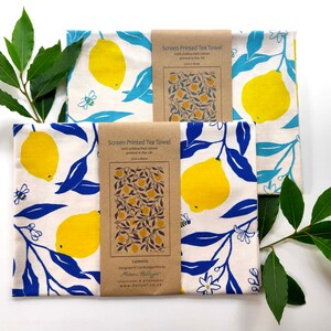 May include: Two white tea towels with a lemon and leaf design. The towels are printed with blue leaves and yellow lemons. The text on the brown label reads "Screen Printed Tea Towel" and "Lemons".