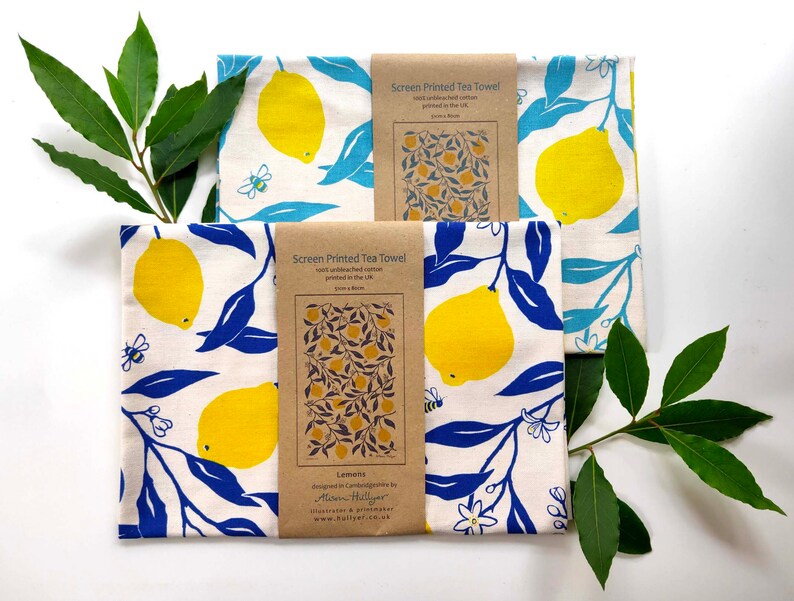 May include: Two white tea towels with a lemon and leaf design. The towels are printed with blue leaves and yellow lemons. The text on the brown label reads "Screen Printed Tea Towel" and "Lemons".