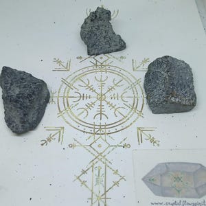 Op de afbeelding: Drie ruwe, donkergrijze stenen zijn gerangschikt rond een goudkleurig cirkelvormig geometrisch symbool dat op een wit oppervlak is gedrukt. Een kleine vierkante kaart met een kristalillustratie en de tekst www.crystalflowspirit is aanwezig.