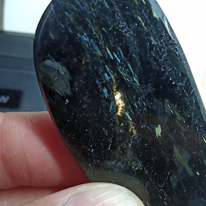 Pode incluir: Uma pedra escura polida com inclusões iridescentes azuis, verdes e douradas. A pedra tem uma forma lisa e curva e um acabamento brilhante, refletindo a luz para criar um efeito cintilante. A cor geral é preto profundo.