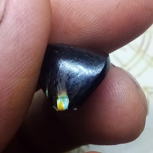 Op de afbeelding: Een gepolijste, donkergekleurde edelsteen met een glad, afgerond oppervlak. De steen vertoont aan één kant een duidelijke, iriserende flits van geel en blauw licht, wat contrasteert met de diepe, donkere basiskleur van het mineraalexemplaar.
