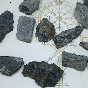 Op de afbeelding: Een verzameling onregelmatige, donkergrijze en zwarte mineraalfragmenten met een ruw, getextureerd oppervlak. De stenen liggen verspreid over een wit oppervlak met een goudkleurig, cirkelvormig geometrisch symbool met fijn lijnwerk.