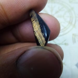 Op de afbeelding: Een gepolijst edelsteenfragment met een duidelijke kleurscheiding tussen een diepzwart, massief gedeelte en een goudbruin gedeelte met zichtbare lineaire, vezelige insluitingen die een chatoyant effect op het oppervlak creëren.