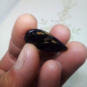 Op de afbeelding: Een donker, druppelvormig object met gouden vlekken, vastgehouden in een hand. Het object is glad en gepolijst en reflecteert licht. De achtergrond is een vage, gebroken witte ondergrond met vage markeringen. De foto is gemaakt met een Blackview BV4900 Al Quad camera.