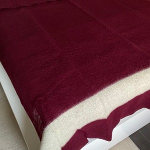 Puede incluir: Una manta burdeos con una franja crema en la parte superior, sobre un marco de cama blanco. La textura de la manta parece suave y afelpada. Adecuada como colcha o manta, mide aproximadamente 200 cm x 150 cm.