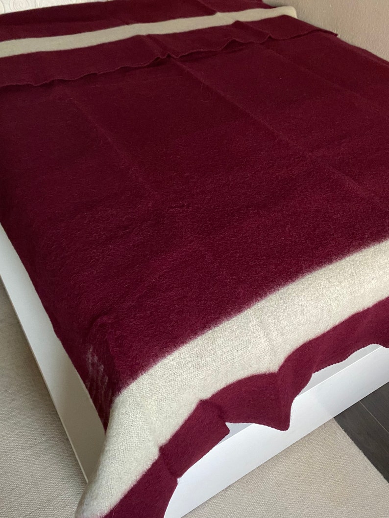 Puede incluir: Una manta burdeos con una franja crema en la parte superior, sobre un marco de cama blanco. La textura de la manta parece suave y afelpada. Adecuada como colcha o manta, mide aproximadamente 200 cm x 150 cm.