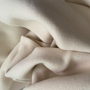 May include: Close-up of a soft, off-white fabric with a textured surface. The fabric is draped in gentle folds, creating shadows and highlights. The material appears to be thick and warm, possibly wool or a similar textile.