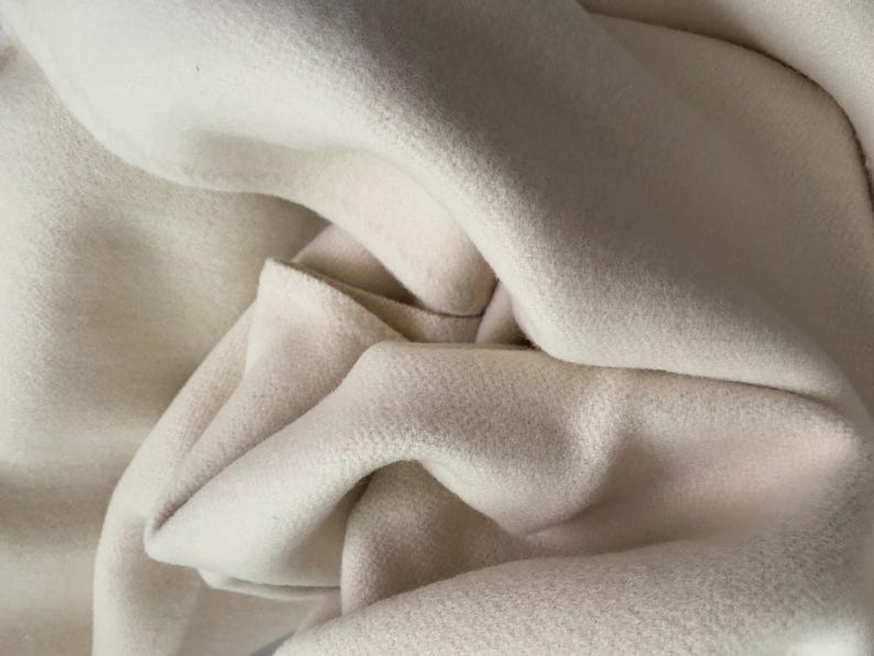 May include: Close-up of a soft, off-white fabric with a textured surface. The fabric is draped in gentle folds, creating shadows and highlights. The material appears to be thick and warm, possibly wool or a similar textile.