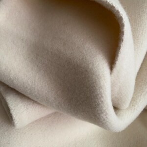 May include: Close-up of a soft, off-white fabric. The material is thick and appears to be wool or fleece. The fabric is folded over itself, showing the texture and depth of the material. The edges are slightly frayed.