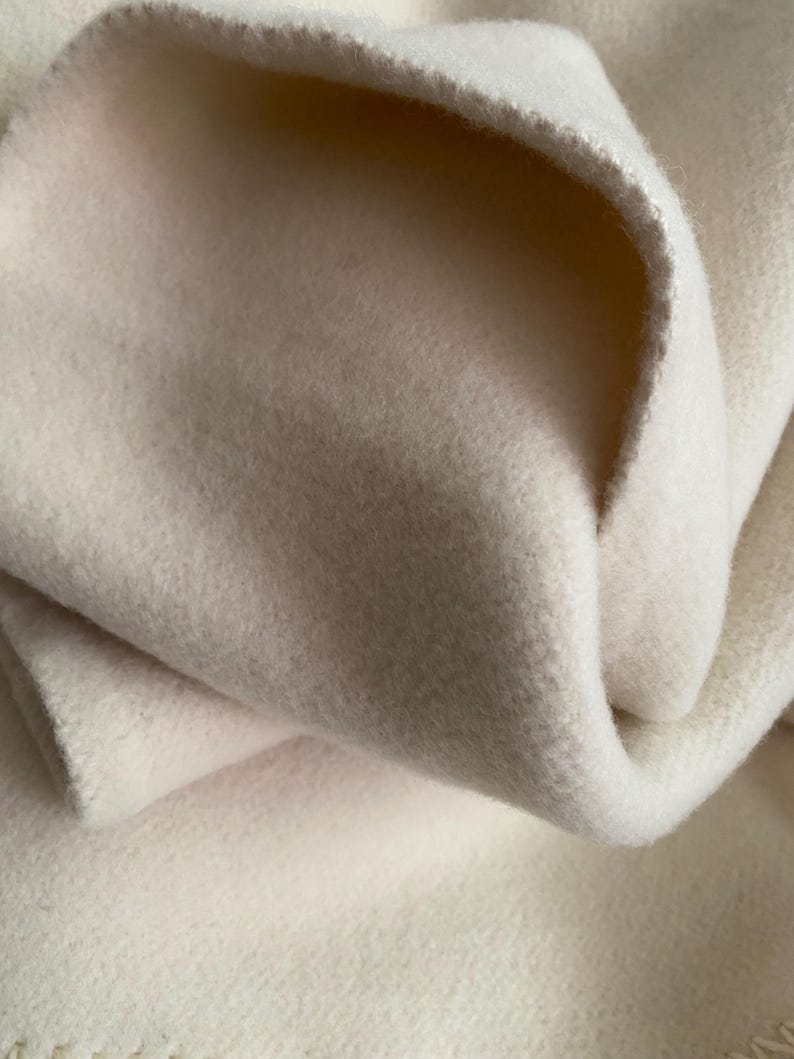 May include: Close-up of a soft, off-white fabric. The material is thick and appears to be wool or fleece. The fabric is folded over itself, showing the texture and depth of the material. The edges are slightly frayed.