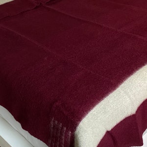 Puede incluir: Una manta burdeos y crema sobre una cama. La manta tiene una ancha franja crema a lo largo de los bordes superior e inferior. El resto de la manta es de color burdeos liso. La manta est&aacute; hecha de un material suave y texturizado.