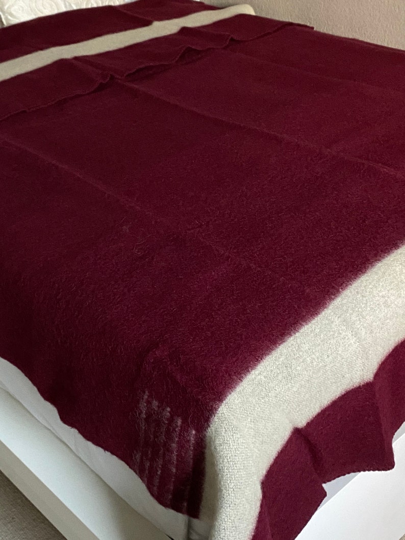 Puede incluir: Una manta burdeos y crema sobre una cama. La manta tiene una ancha franja crema a lo largo de los bordes superior e inferior. El resto de la manta es de color burdeos liso. La manta est&aacute; hecha de un material suave y texturizado.