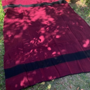 Peut inclure: Une couverture bordeaux avec une large bande noire près des bords supérieur et inférieur. La couverture est étalée sur une surface herbeuse, avec des ombres de feuillage. La couverture semble être faite d'une matière douce et chaude.