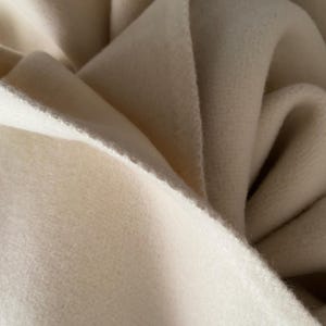 May include: Close-up of a soft, off-white fabric with a textured surface. The fabric is folded and draped, creating shadows and highlights. The material appears to be thick and warm, possibly wool or fleece.