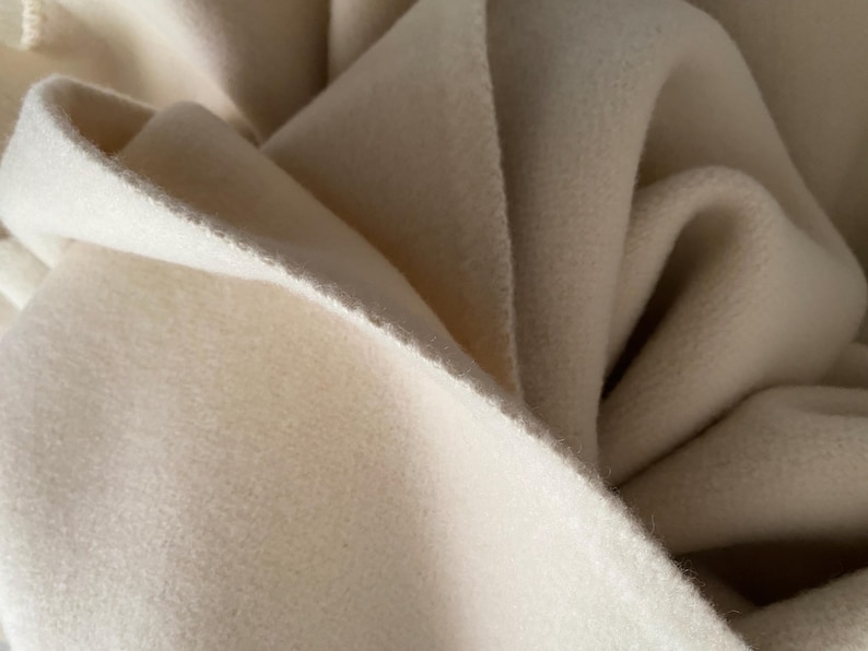 May include: Close-up of a soft, off-white fabric with a textured surface. The fabric is folded and draped, creating shadows and highlights. The material appears to be thick and warm, possibly wool or fleece.