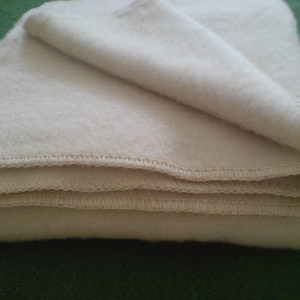 May include: Folded cream-coloured blanket with visible stitching along the edges. The blanket appears soft and plush, with a folded corner revealing the layers. The background is a solid green colour.
