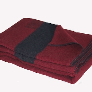 Op de afbeelding: Gevouwen bordeauxrode deken met een brede zwarte streep. De deken is gemaakt van een dik materiaal en heeft een gestikte rand. De gevouwen deken ligt op een witte achtergrond.