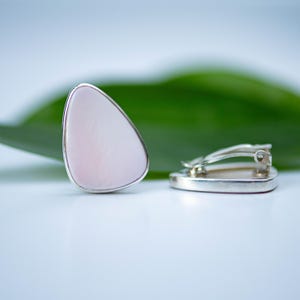 May include: A pair of teardrop-shaped clip-on earrings featuring smooth, pale pink stone cabochons set in polished silver-toned metal frames. One earring is positioned upright to show the face, while the other lies flat to reveal the metal clip mechanism.