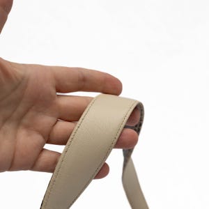 May include: Close-up of a beige leather strap with a stitched edge. The strap is held by a hand against a white background. The strap appears to be part of a bag or purse.