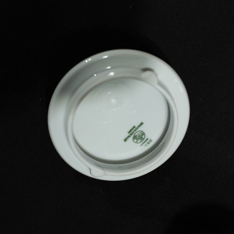 May include: A white porcelain ashtray with a circular design. The ashtray has a recessed center and a small notch for resting cigarettes. The bottom features green text and a logo that reads "LORE BAVARIA REUTTER GERMANY LESIE".