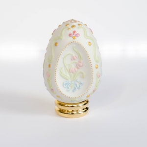 以下が含まれることがあります： 金色のアクセントと花柄が施された白い磁器の卵。卵は金色の台座の上に置かれています。