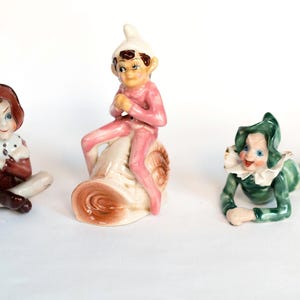 May include: Three vintage ceramic figurines. One figure is seated with crossed arms, wearing a brown outfit with a white collar. Another figure is seated on a snail, wearing a pink outfit. The third figure is lying down, wearing a green outfit.
