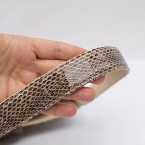 May include: A close-up of a snakeskin-patterned belt. The belt features a beige and brown scale pattern on the exterior and a cream-colored interior. The belt is held in a hand against a white background.