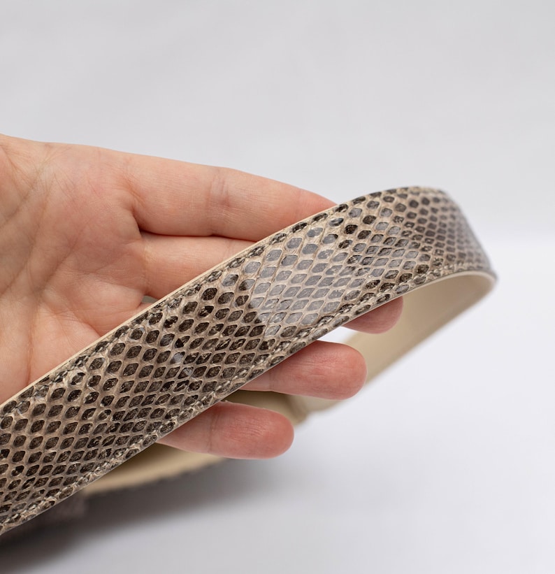 May include: A close-up of a snakeskin-patterned belt. The belt features a beige and brown scale pattern on the exterior and a cream-colored interior. The belt is held in a hand against a white background.