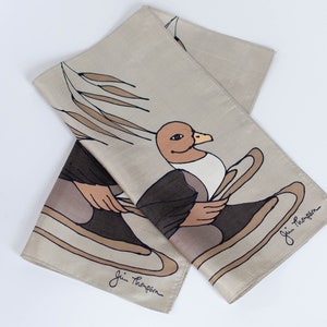 May include: A beige silk scarf featuring a cartoon duck design. The duck is illustrated in brown, black, and beige, holding oars. The scarf is folded, with the artist's signature visible in the corner.
