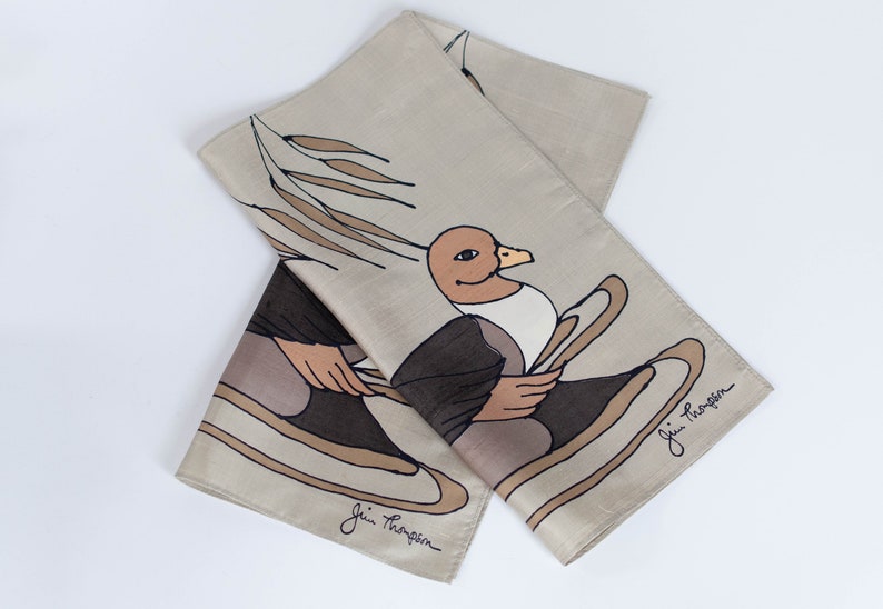 May include: A beige silk scarf with a whimsical duck design. The duck is depicted in shades of brown, black, and beige, holding oars. The scarf has a folded design with the artist's signature in the corner.