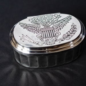 May include: A silver oval-shaped box with a hinged lid. The lid features an engraved image of the Great Seal of the United States, which includes an eagle with outstretched wings, a shield with thirteen stripes, and an olive branch and arrows.