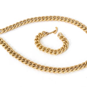May include: A gold-coloured chain necklace and matching bracelet set. The necklace and bracelet feature a chunky curb chain design. The jewellery is displayed on a white background.