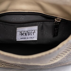 May include: Interior view of a beige and gray snakeskin-patterned handbag. The inside is lined with a dark gray fabric. A label reads "MONICA MAGLI MADE IN ITALY". A zipper pull is visible.