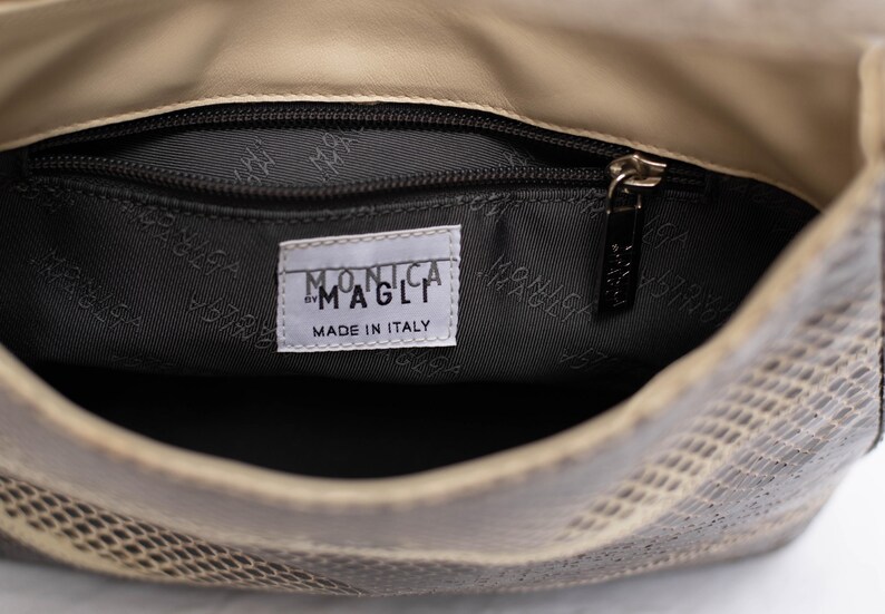 May include: Interior view of a beige and gray snakeskin-patterned handbag. The inside is lined with a dark gray fabric. A label reads "MONICA MAGLI MADE IN ITALY". A zipper pull is visible.